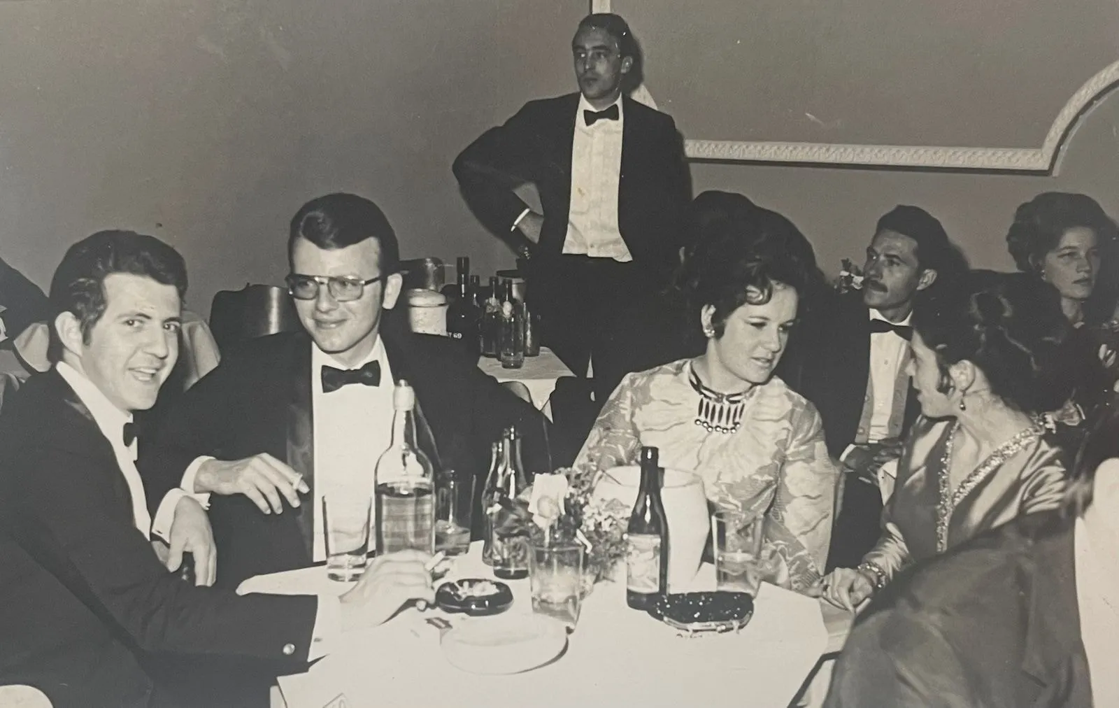 A mesa do baile, com quatro pessoas em trajes de gala sentadas entre garrafas e taças, e um homem de smoking em pé ao fundo