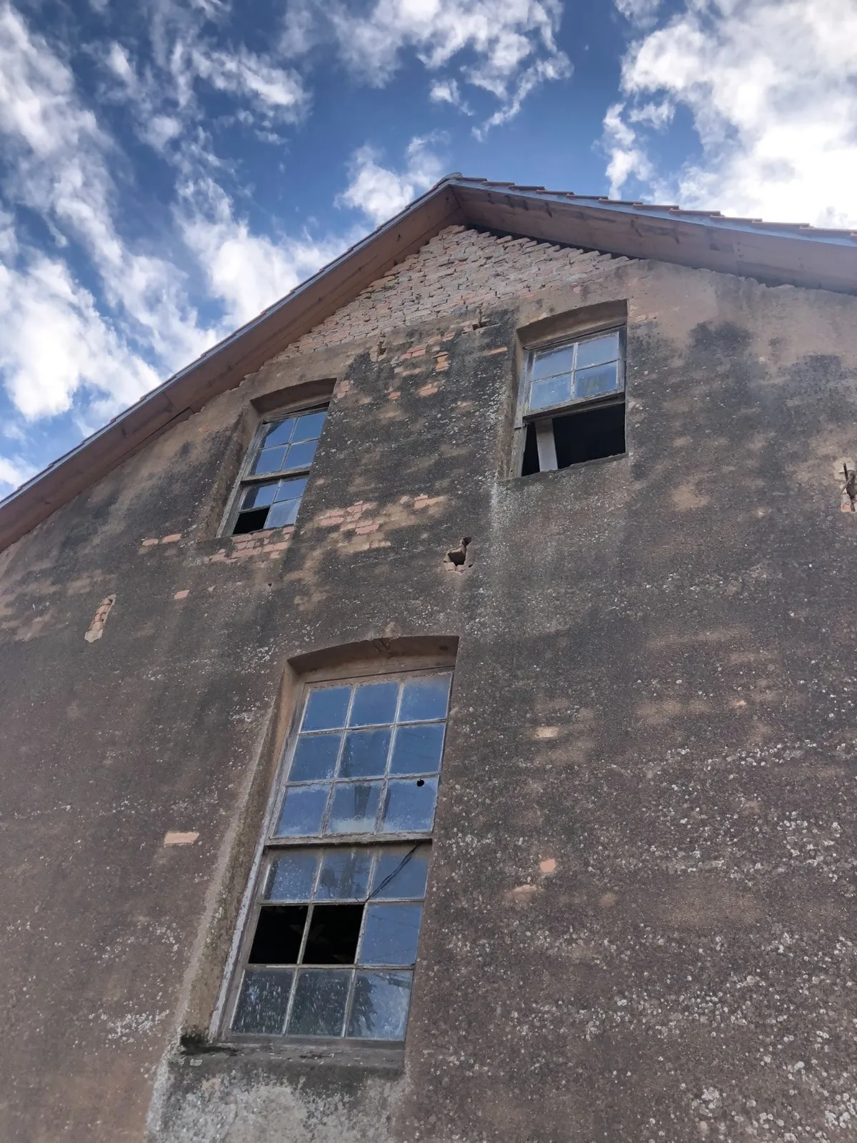Detalhe da mesma parede vista de baixo, com reboco caído mostrando os tijolos e vidros quebrados nas janelas